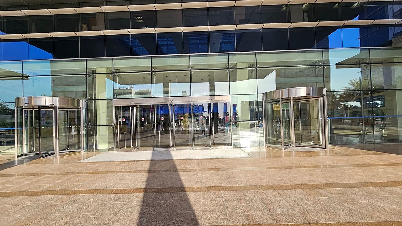 A modern building entrance with large glass doors and windows. Two revolving doors are on the left and right, framed in metallic silver. The central section features multiple glass sliding doors. The floor is tiled in a pattern of light and dark brown. Reflections of the surrounding area, including trees and other structures, are visible in the glass. The ceiling inside is white with recessed lighting.
