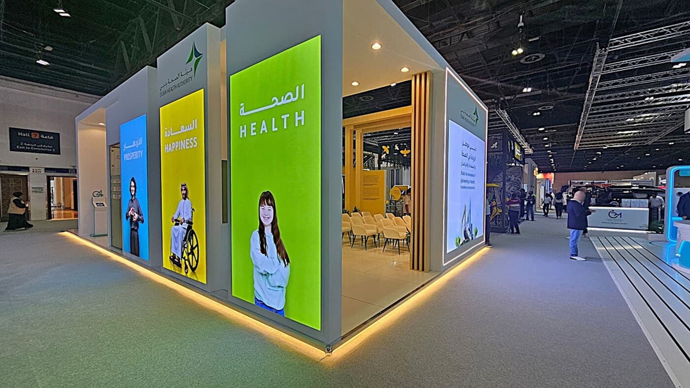 A modern exhibition booth with large vertical screens displaying the words "Prosperity," "Happiness," and "Health" in both English and Arabic. The screens feature images of diverse individuals. Inside the booth, there are rows of white chairs. The floor is carpeted in grey, and the ceiling has exposed beams with spotlights. To the right, people are walking through the exhibition hall, and there are various other booths visible in the background.