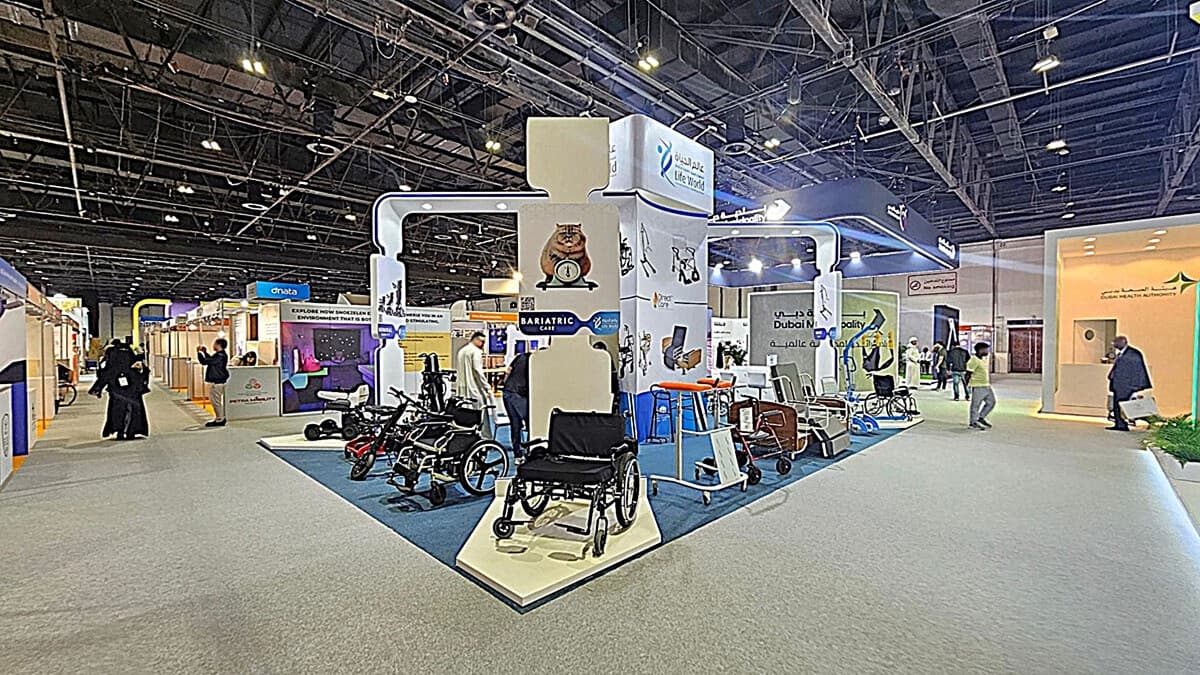 A trade show booth with various mobility aids, including wheelchairs and walkers, displayed on a blue carpeted platform. The booth features signage for bariatric care and is surrounded by other exhibition stands. People are walking around, engaging with the displays. The ceiling is high with exposed beams and lighting fixtures. The floor is covered with grey carpet.
