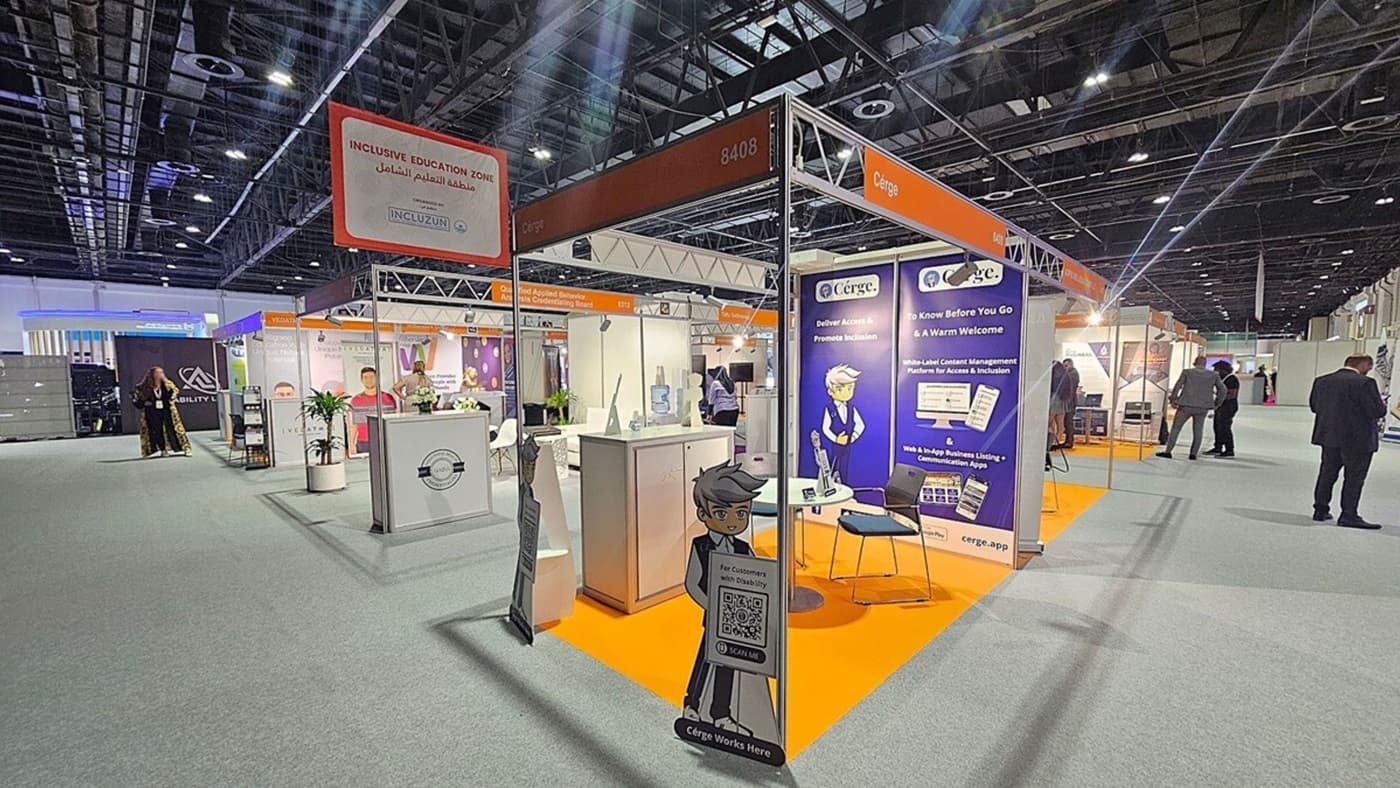 A trade show booth with a sign reading "Inclusive Education Zone" in both English and Arabic. The booth features a display for "Cérge," with promotional materials and a cartoon character holding a QR code sign. The floor is carpeted in grey, and the booth area is marked with an orange carpet. Several people are walking around the exhibition hall, which has a high ceiling with exposed beams and lighting fixtures. Other booths are visible in the background.