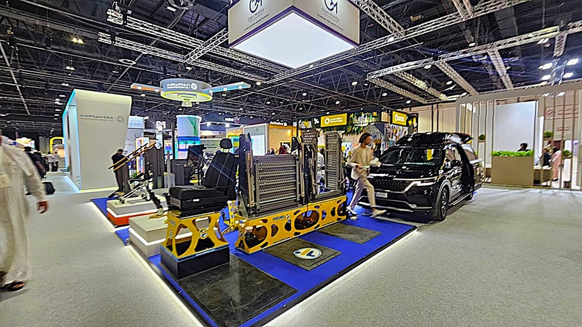 A trade show floor with various exhibits. On the left, there are display stands with equipment and a person walking by. In the centre, a platform with mechanical components and seating is showcased on a blue mat. On the right, a black vehicle with open doors is displayed. The background features booths with signage and people interacting. The ceiling has a grid of lights and metal trusses. The floor is carpeted in grey.