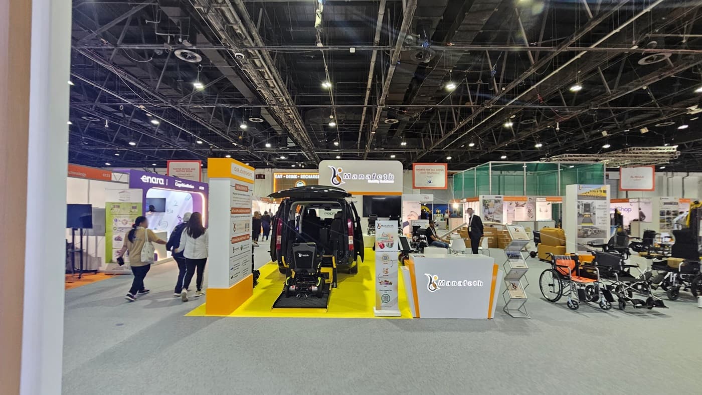 A trade show booth with a black vehicle in the centre, featuring an accessibility ramp extending onto a bright yellow platform. The booth is branded with "Manafeth" and includes informational displays. On the right, there are various mobility aids like wheelchairs. In the background, other booths are visible with people walking around. The ceiling is industrial with exposed beams and lighting fixtures. The floor is carpeted in grey.