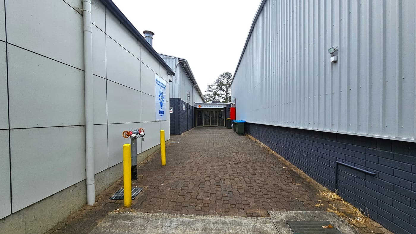 In the middle is an alleyway featuring a brown, brick pathway that is uneven in some areas. The Recreation Centre is on the right, a building with corrugated walls. On the left is another grey building for the Football Club. Yellow bollards are on the left, along with a fire hydrant. At the end is the entrance to Adelaide Hills Recreation Centre.