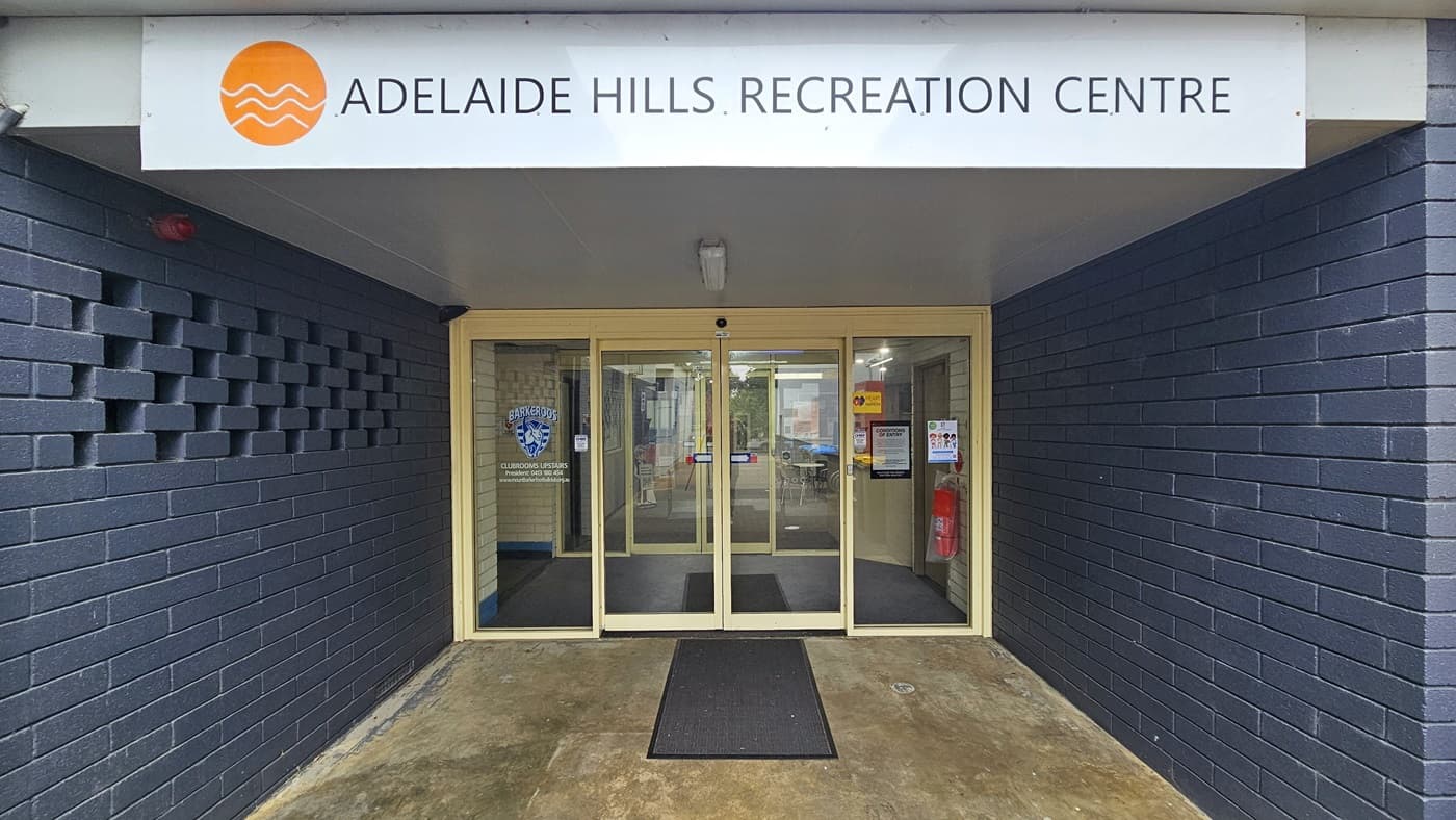 The entrance is enclosed by dark blue brick walls. The door consists of two sets of automatic, glass sliding doors with stickers and posters on the wall, including a fire extinguisher. A non-slippery mat is placed on the doorstep. A distinctive sign, "Adelaide Hills Recreation Centre," is positioned on top near the ceiling.