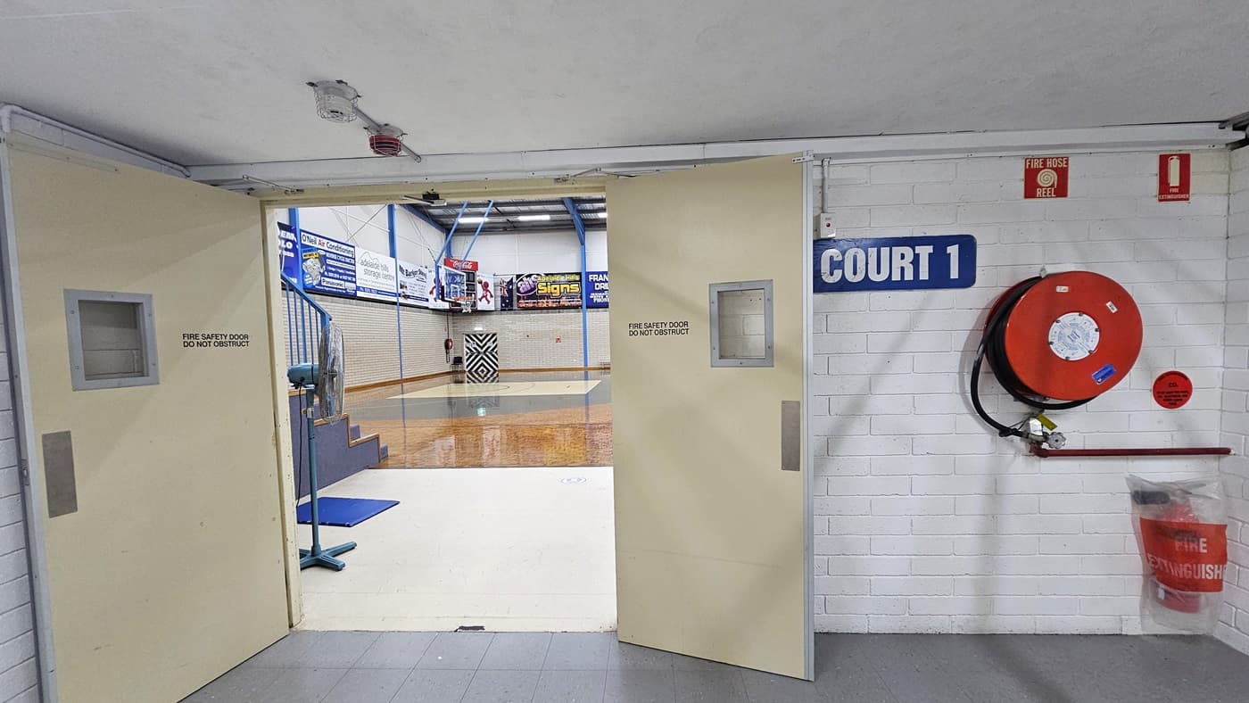 The entrance door, painted in white, is made of wood. To the right, a sign reads "Court 1”. On the left wall, a fire extinguisher and a fire hose reel are mounted. Inside, a section of the court is visible, showcasing the timber floor, exposed beams, and signages.