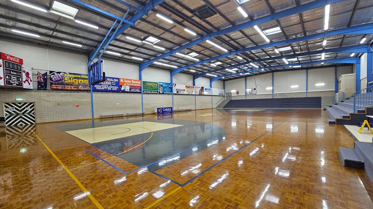 Court 1 has a polished timber parquet floor with game markings. Tiered seating on the right and middle provides spectator views. Walls around the courts display advertising materials. The ceiling features exposed steel beams, fluorescent lights, and insulation foams. A digital screen is in the left corner, and below is an emergency exit door. The basketball boards are placed on both sides.