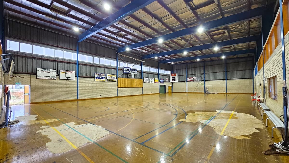 Court 2 features a sturdy concrete floor with game markings, showing signs of worn-out paint in some areas. On the left, bench chairs are available for seating. The court is enclosed by a white brick wall with boards and posters on the right. The ceiling is equipped with exposed steel beams, fluorescent lights, and insulation foams. Basketball boards are placed on both sides.