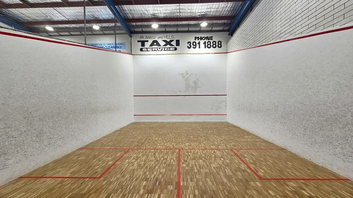 The Squash Court is visible from the viewing deck, enclosed by metal railings with a net. The court features a timber floor marked with vibrant red lines. Advertisements adorn the white walls, and the high ceiling is illuminated by fluorescent lights and equipped with insulation forms.