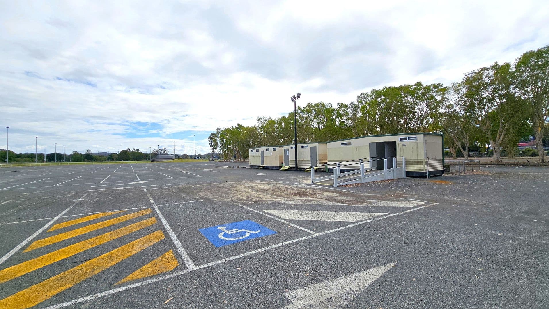 A large, mostly empty car park with marked parking spaces, including a blue disabled parking spot in the foreground. Yellow diagonal lines are painted on the ground nearby. On the right, several portable buildings are lined up, with a ramp leading to one of them. Trees are visible in the background, and a few streetlights are scattered throughout the area. The sky is overcast.