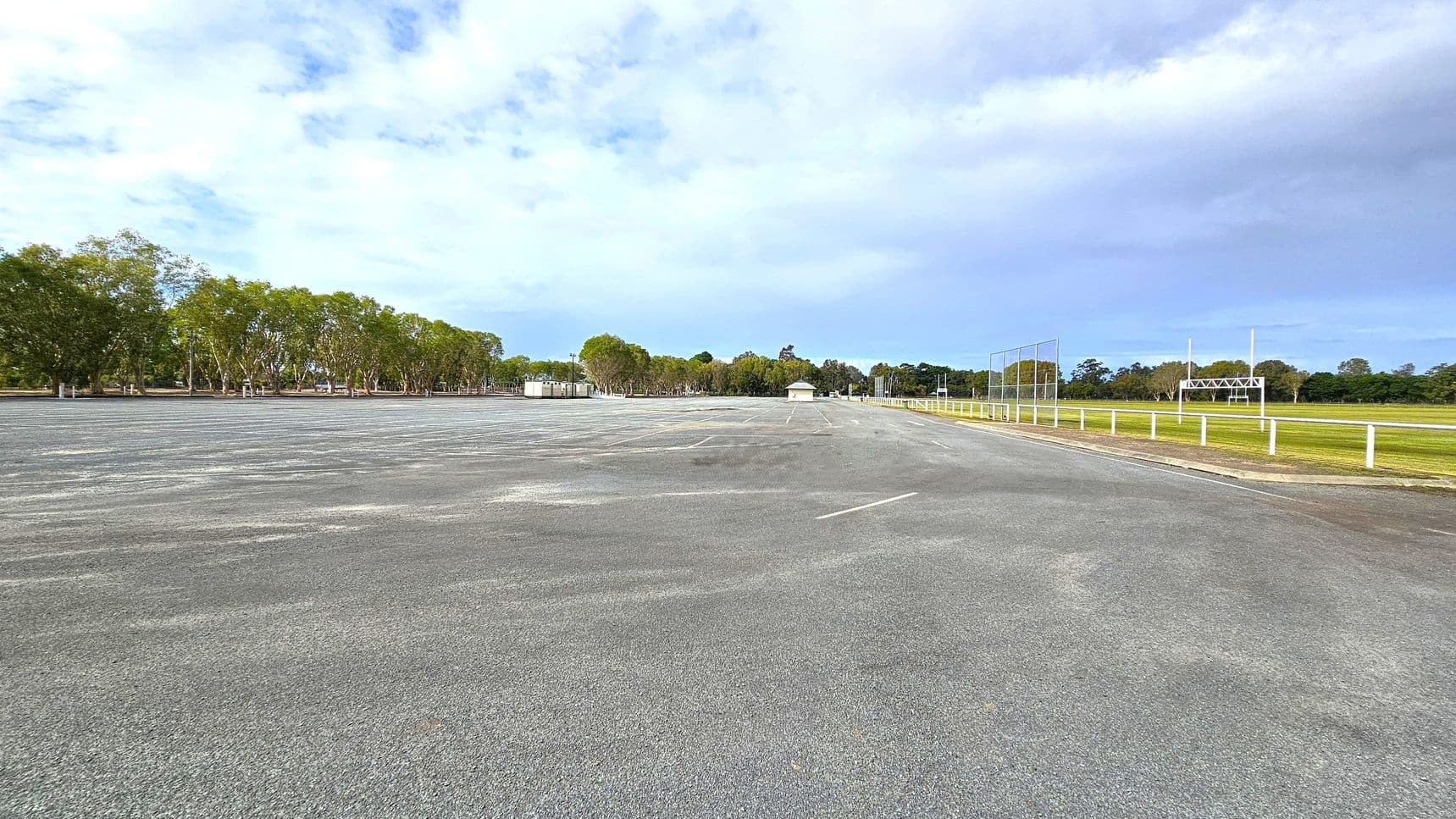 A large, empty car park with a grey asphalt surface stretches across the foreground. On the left, a row of trees lines the edge, providing a natural border. In the middle, there are small structures, possibly sheds or kiosks. On the right, a grassy field is enclosed by a white fence, with a scoreboard and goalposts visible. The background features more trees, creating a green backdrop. The sky is overcast, casting a soft light over the scene.
