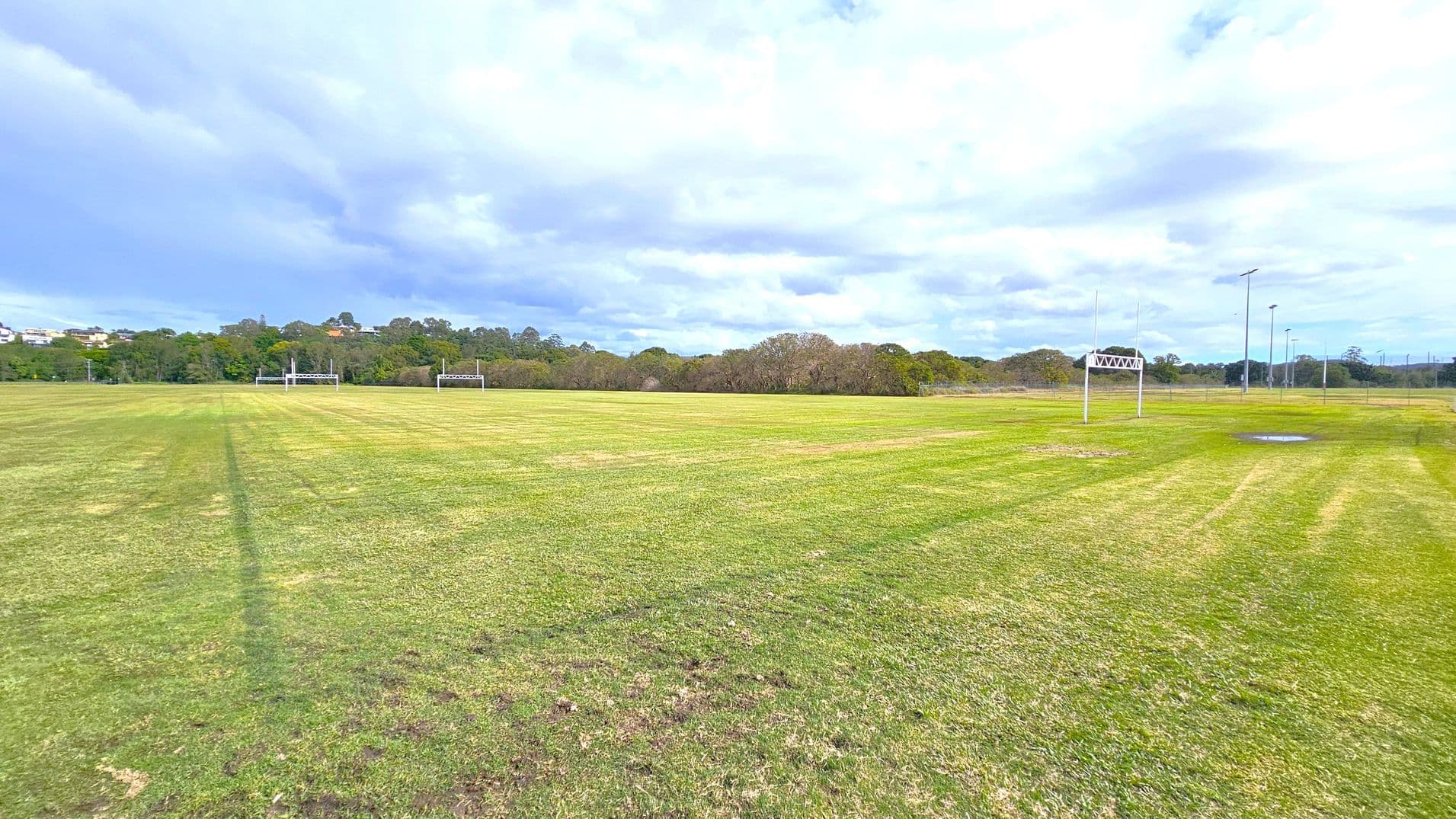 A large grassy field with rugby goalposts on the left and right sides. The grass is a mix of green and brown patches. In the background, there are trees and some distant buildings partially visible. Several tall floodlights are positioned on the right side of the field. The sky is overcast with a mix of light and dark clouds. A small puddle of water is visible on the right side of the field.