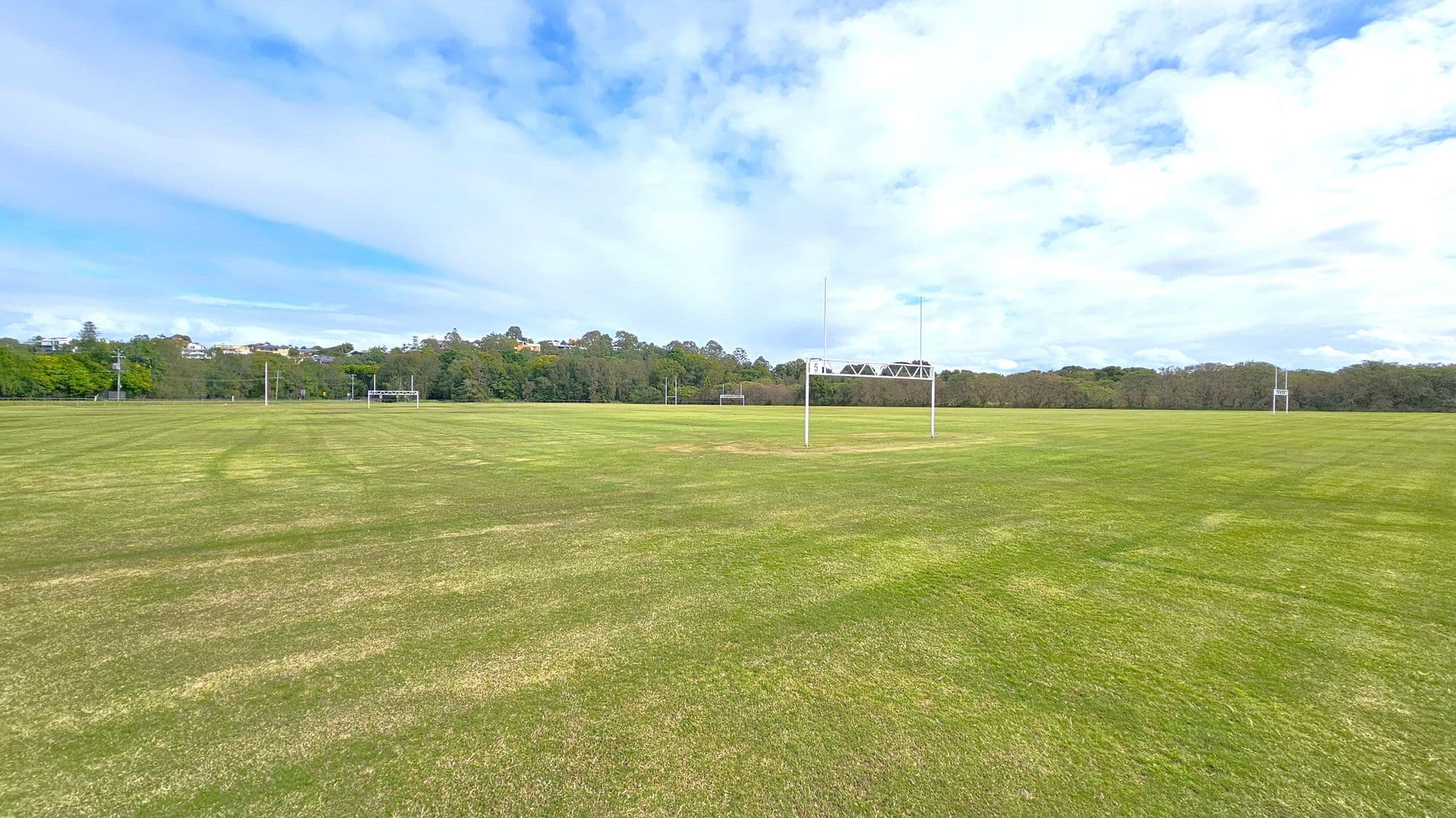 A large, open grassy field with rugby goalposts in the centre and to the right. The grass is a mix of green and brown patches. In the background, there are trees and a few buildings visible on a slight hill. The sky is mostly cloudy with some blue patches.