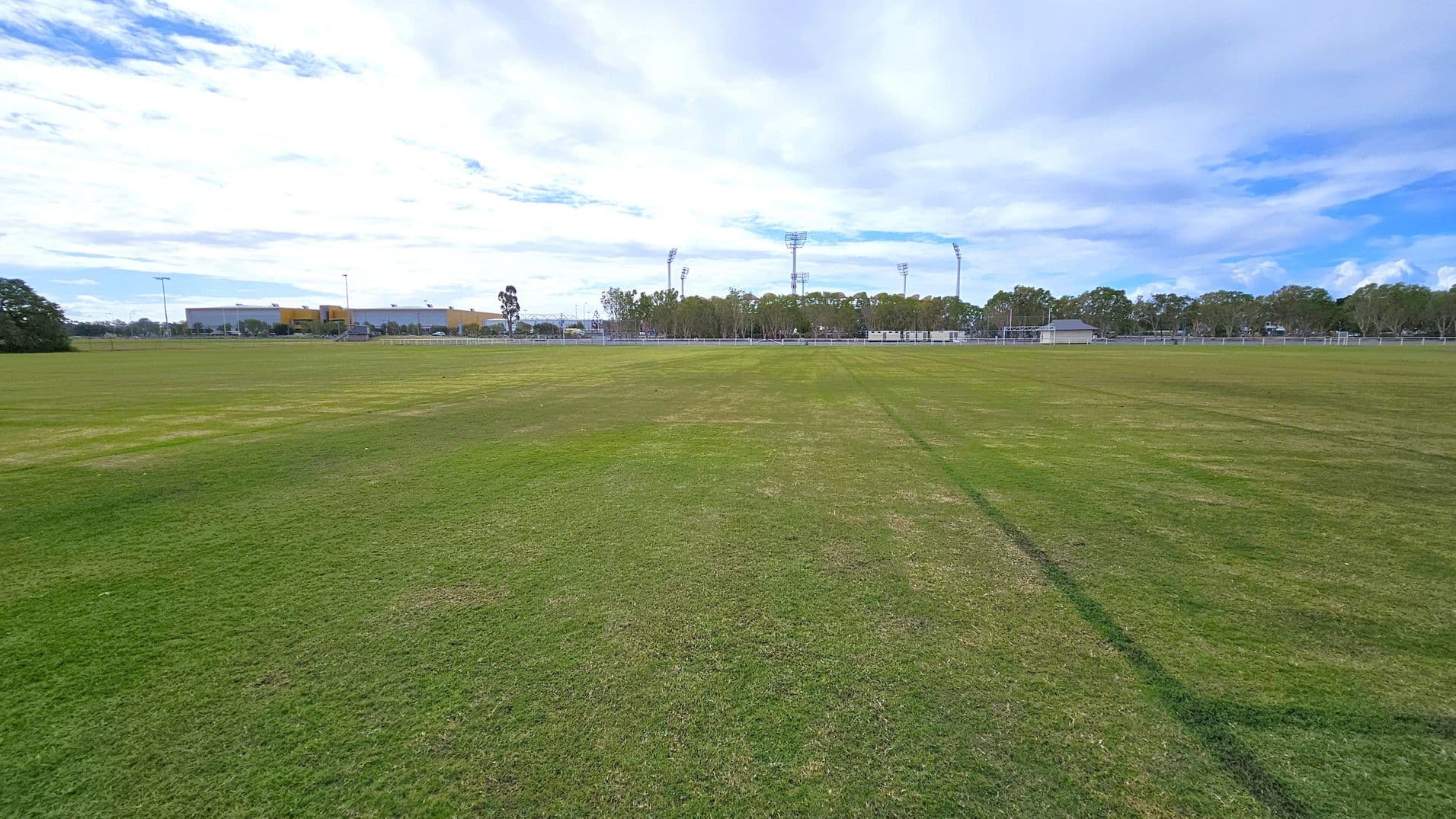 A wide, open grassy field stretches across the foreground, with patches of varying green shades. In the background, there are several tall floodlights and a line of trees. To the left, a large building with a yellow and grey facade is visible, and a few smaller structures are scattered to the right. The sky is mostly cloudy, casting a soft light over the scene.