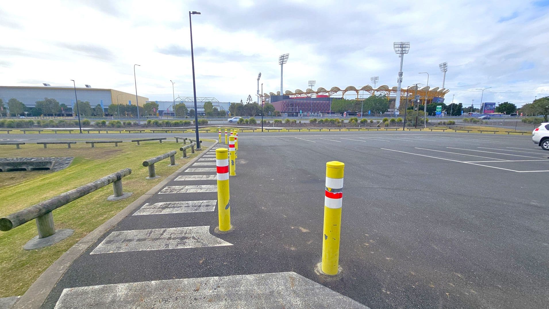A parking area with yellow bollards lined up along a pedestrian crossing. The ground is asphalt with white painted lines. On the left, there is a grassy area with wooden barriers. In the background, a large building with a curved roof and tall floodlights is visible, likely a stadium. A few trees and shrubs are scattered around, and a white car is parked on the right side. The sky is overcast.