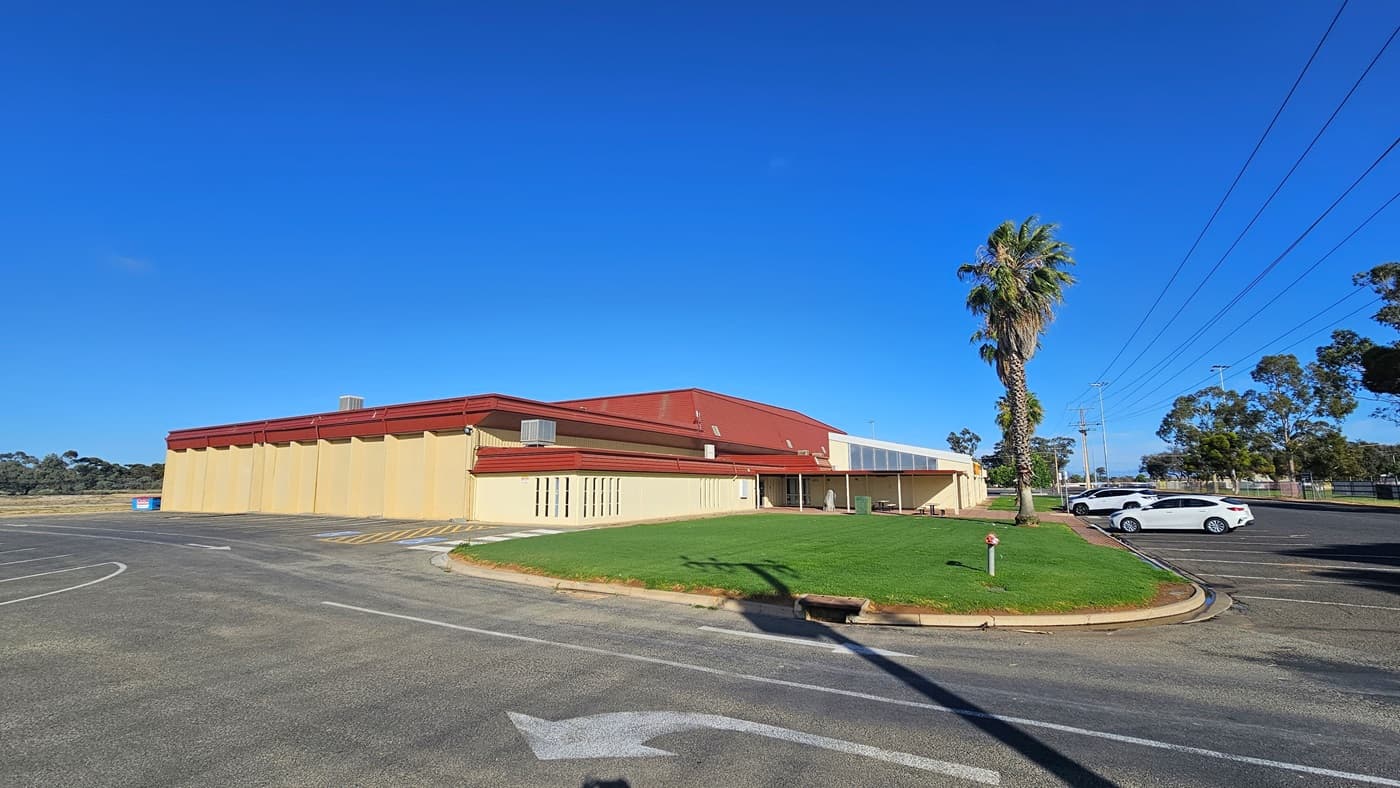A cream industrial building with red corrugated roofing. At the front, manicured grass and palm trees adorn the landscape. Two white cars are parked in the car park area in front of the building. A garden light post is visible on the grass. Further ahead, trees can be seen. On the right side of the building, additional car parking is available, including accessible parking marked with blue wheelchairs and yellow diagonal lines.