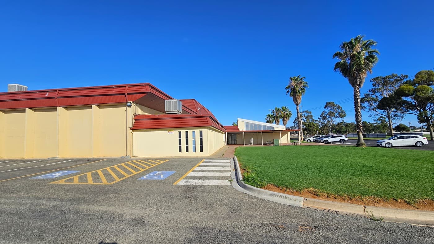 To the left stands a cream industrial building with red corrugated roofing. There is accessible parking marked with blue wheelchairs and yellow diagonal lines. At the front, manicured grass and palm trees adorn the landscape. A few white cars are parked in the car park area in front of the building. Further ahead, trees are visible.