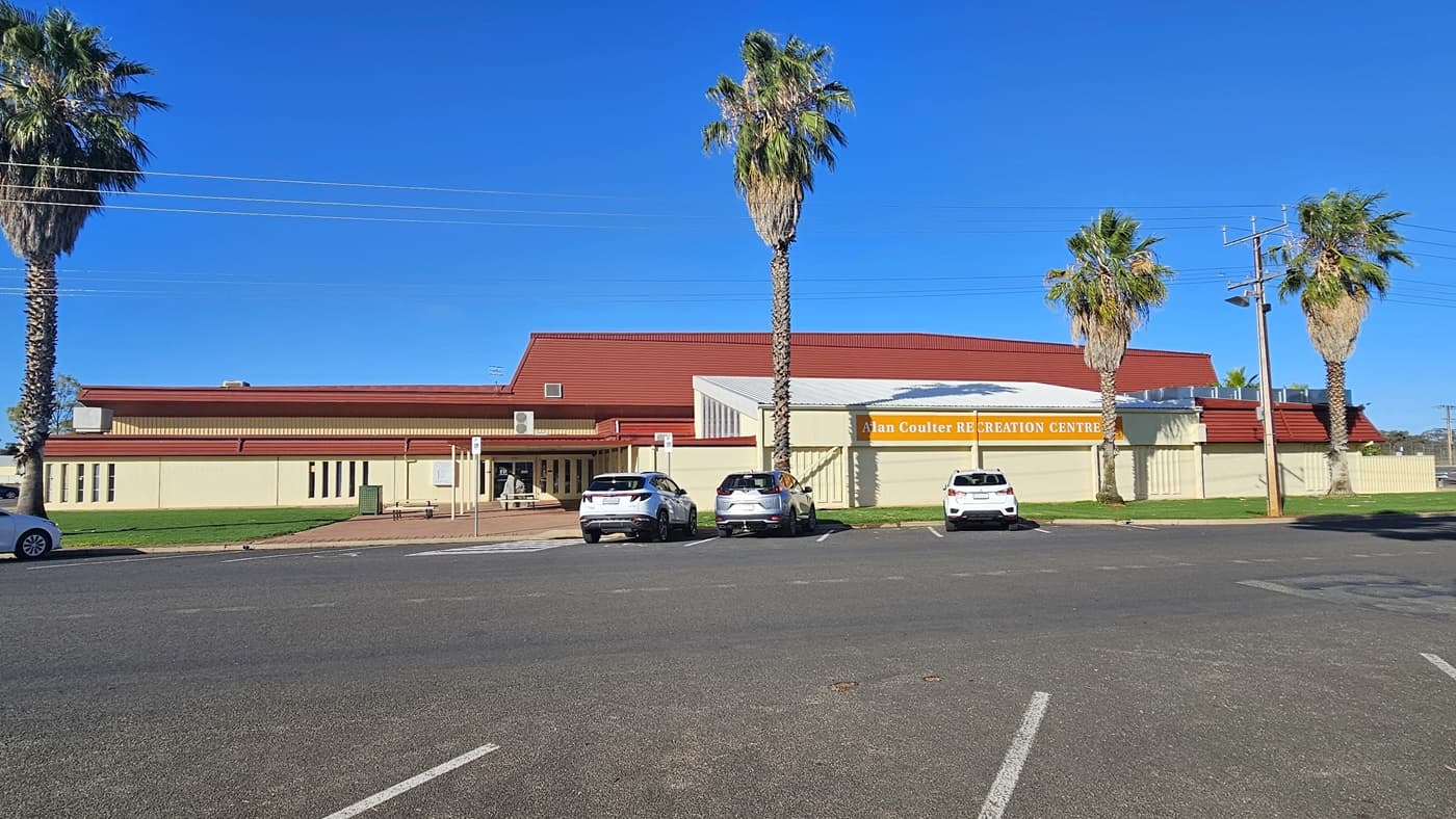 A cream industrial building with red corrugated roofing. In front is a parking area with four cars of various colors and models parked. In the middle, the facility's signage in yellow and white bold letters is visible. Tall palm trees border the parking area. The building's concourse has red bricks and manicured grass.