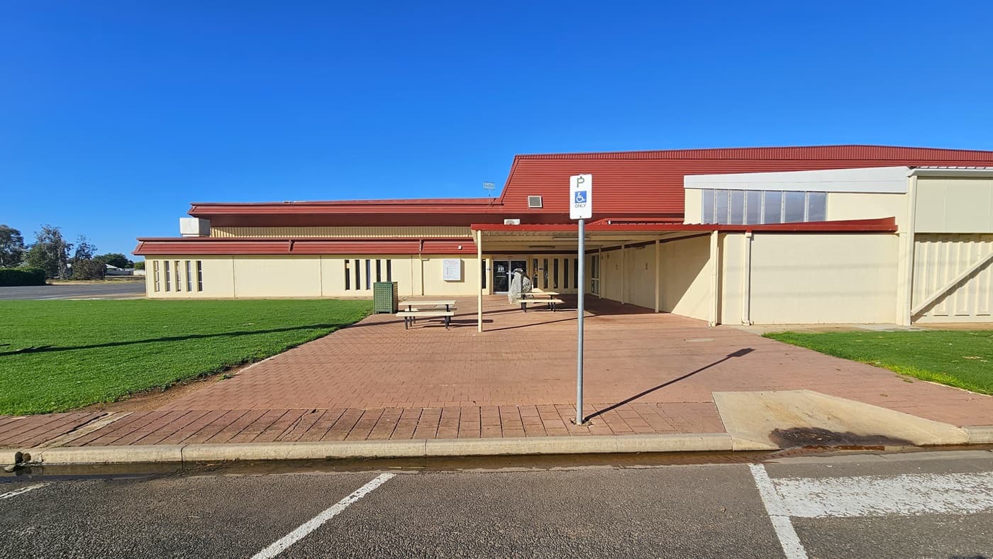 A cream industrial building with red corrugated roofing. The building's concourse has red bricks and manicured grass on both sides. A step-free entrance from the car park, marked by white lines and accessibility signage, is available on the left. Picnic tables are located near the entrance, along with a rubbish bin.
