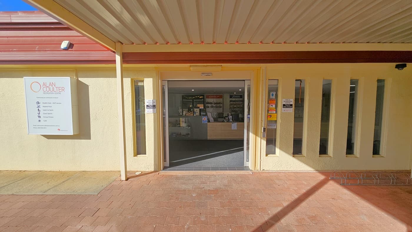 A cream industrial building with red corrugated roofing. The building's concourse is adorned with red bricks. The entrance features an automatic glass door. The left and right walls are a combination of concrete and glass, with a few sticker banners affixed. To the left, the facility signage is displayed, and a bike rack is positioned on the right.
