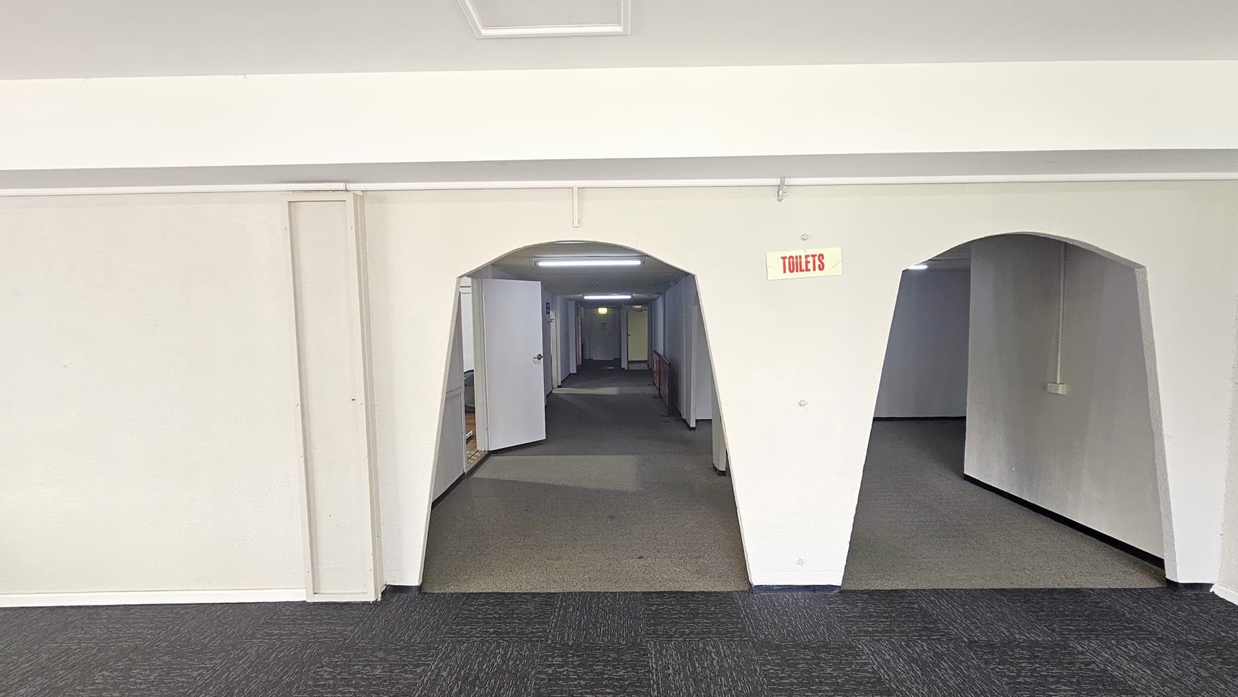 Entrance to the Hydraulic and Cardio Circuit Rooms is through two arches with a "Toilets" sign. The left arch leads to a dimly lit white corridor. A white door opens outward. The walls, ceiling, and floor are all white, with a dark grey carpet.