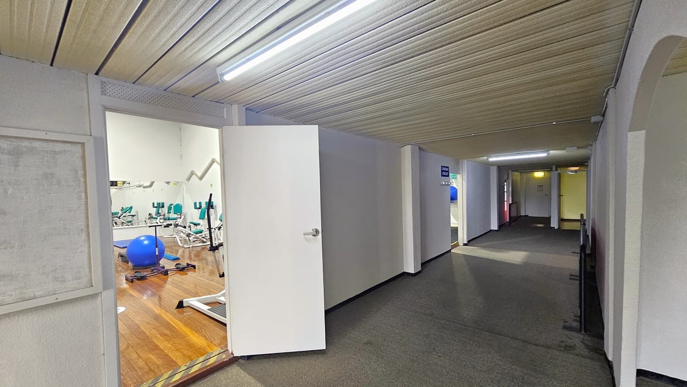 To the left, the outward-opening door reveals the Hydraulic Cardio Room. The room features a small 5 cm stepped entry highlighted in yellow to minimize trip hazards. Inside, resistance machines, including a blue stability ball, are present on a timber floor. The ceiling is painted white with a corrugated design, and the floor has grey carpet. Further ahead is the Circuit room.