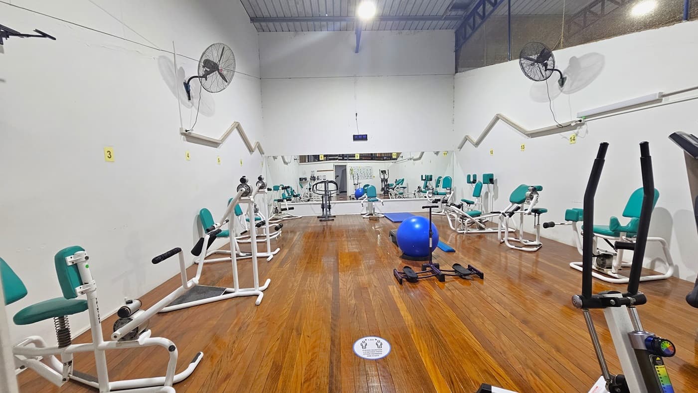 Inside the Hydraulic Cardio Room, resistance machines such as the Abdominal Crunch Machine, Shoulder Press Machine, Calf Machine, and Dip Press machines are arranged. A blue stability ball is placed in the middle. The floor is made of polished timber. Wall fans are mounted on both sides, and the walls are white. The ceiling is white with exposed beams and timber linings.