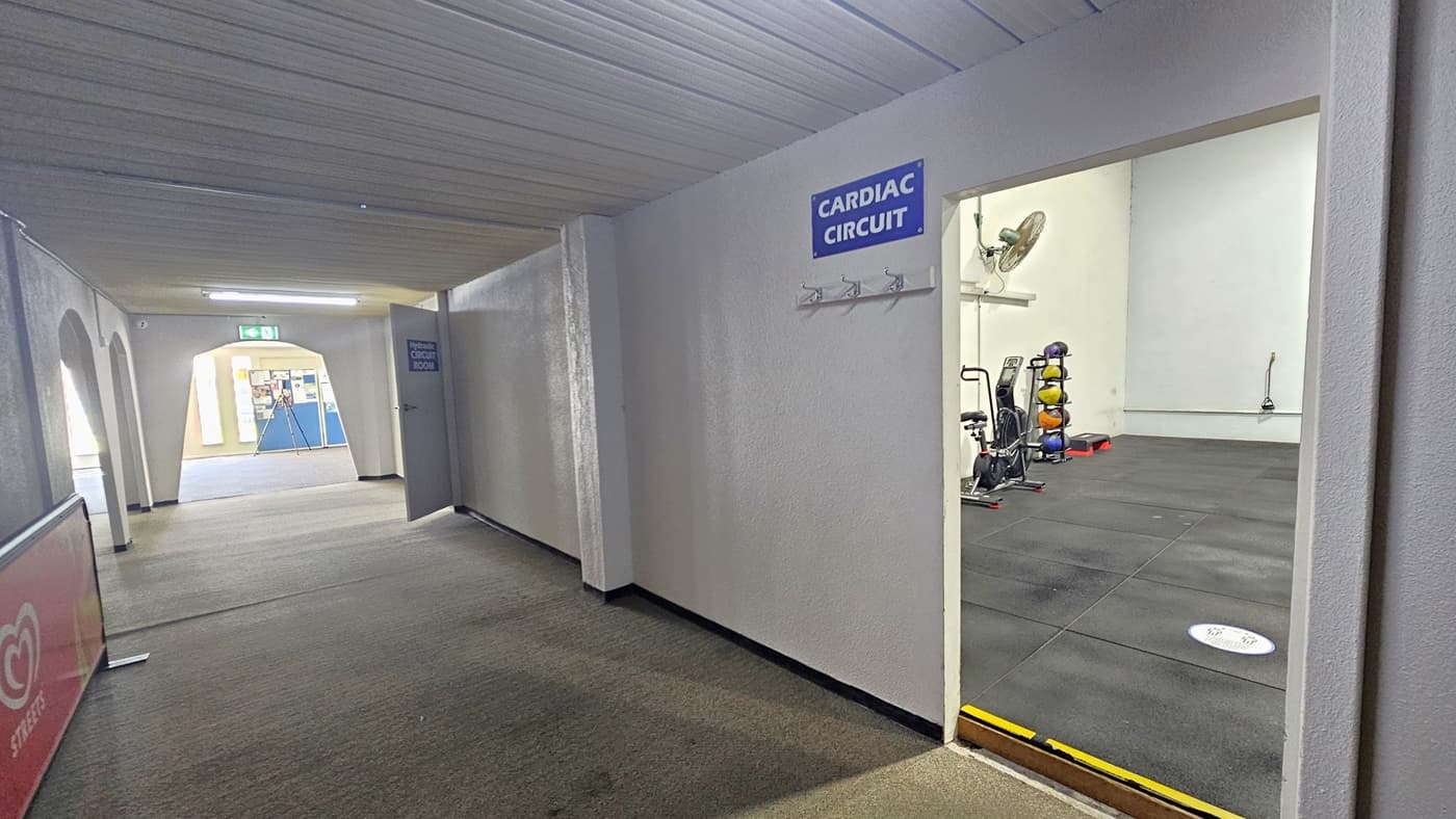 The Circuit Cardiac room on the right features black rubber tiles, wall hooks below blue signage, and a 5 cm yellow-highlighted step entry for safety. Inside, medicine balls are stacked by the left wall, and a stationary bike is present. A wall fan is on the left. Outside, a white ceiling with timber linings and grey carpeted floor is observed. An advertising sign is on the left, and behind the arch, a blue board displays posters.