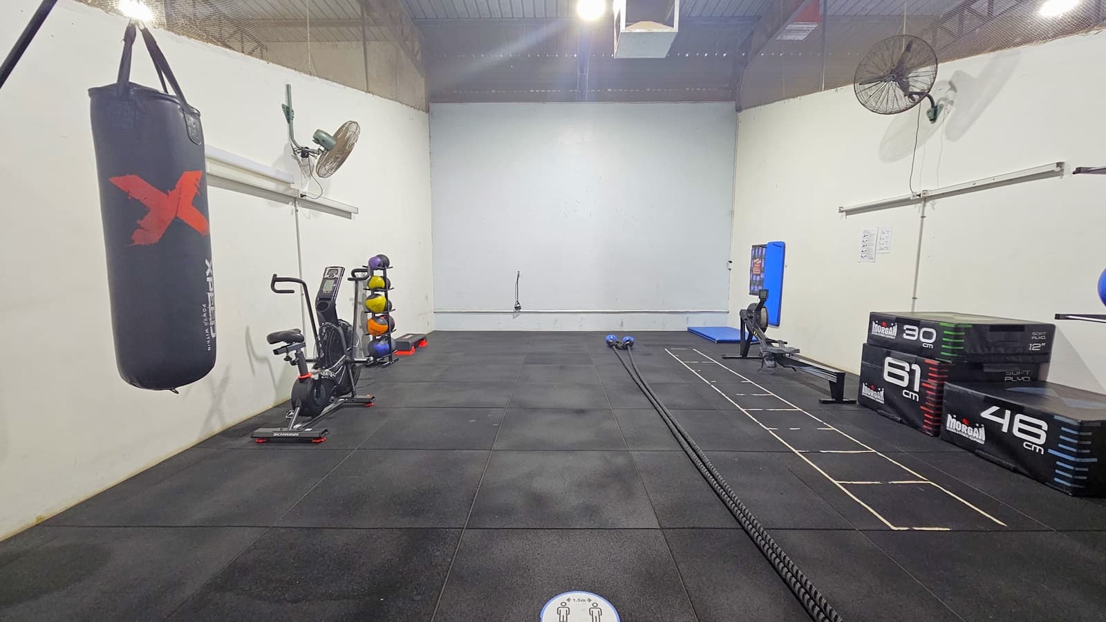 In the Cardiac Circuit Room, stability balls, medicine balls, a punching bag, and a stationary bike are on the left, while a rowing machine, mats, and a long jump rope are on the right. The floor has black rubber mats with white step-like markings. Wall fans are on both sides of the walls, and the ceiling is white with timber linings.