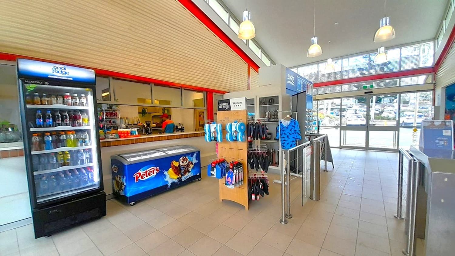 A shop interior with a tiled floor and a high ceiling featuring hanging lights. On the left, there's a refrigerated display filled with drinks and a freezer with "Peters" branding. In the centre, a stand displays various items like swimwear and accessories. The right side has a glass entrance with a view of parked cars outside. The back wall has a counter with snacks and drinks, and a person is visible behind it.