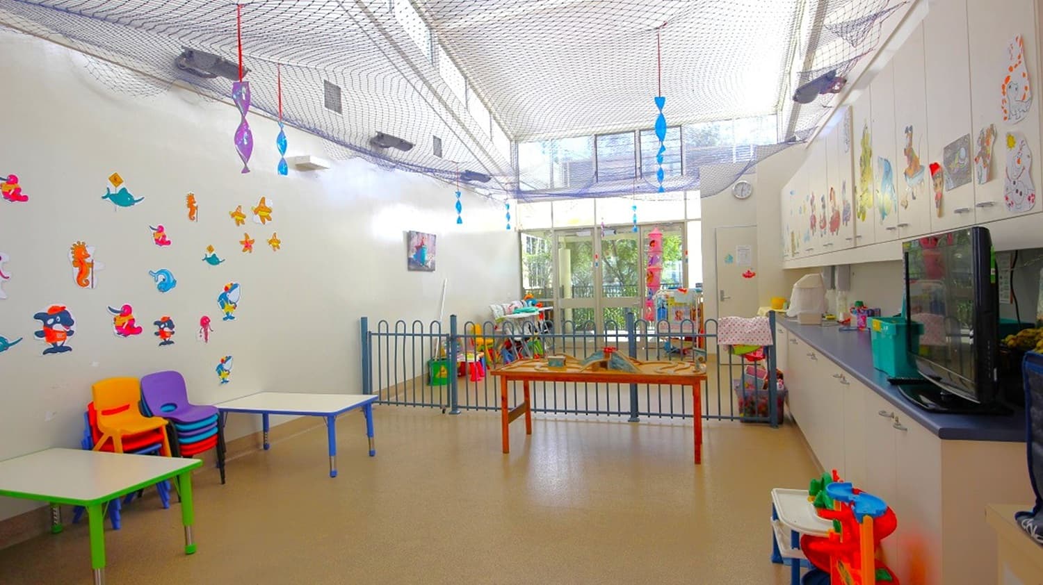 A brightly lit children's playroom with a high ceiling and netting. On the left, colourful chairs and tables are stacked, with cartoon character stickers on the wall. In the centre, a small fenced area contains toys and a table. The right side features a long counter with storage cabinets above and various items on top. The floor is light-coloured, and large windows in the background let in natural light. Hanging decorations are suspended from the ceiling.
