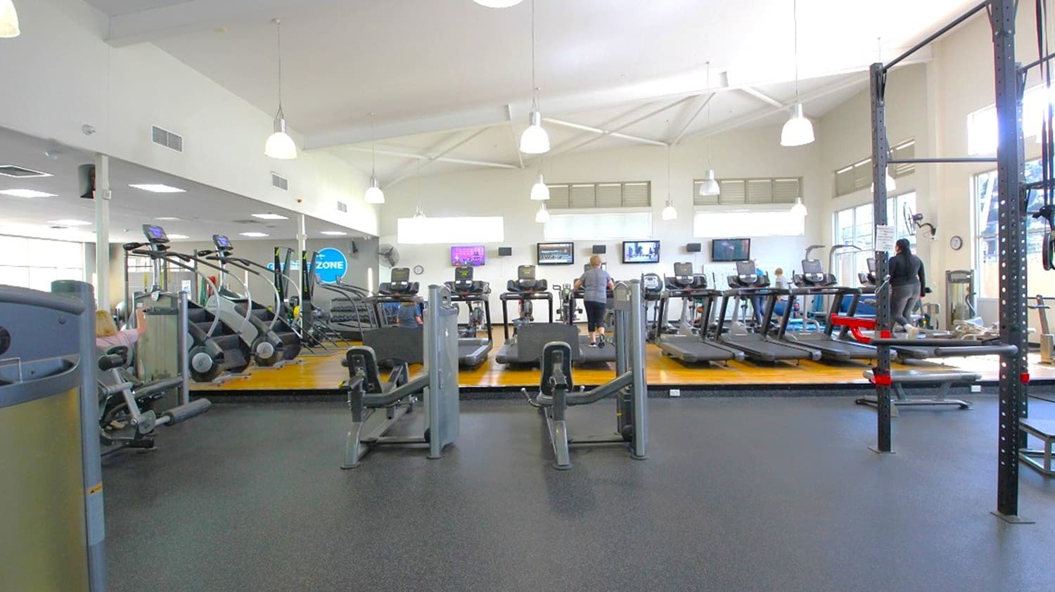 A spacious gym interior with a variety of exercise equipment. In the centre, there are several treadmills and elliptical machines on a raised wooden platform. To the left, more cardio machines are visible. On the right, a person is using a piece of equipment. The ceiling is high with white pendant lights, and the floor is covered with dark rubber mats. Large windows on the right allow natural light to enter, and several wall-mounted TVs are visible in the background.