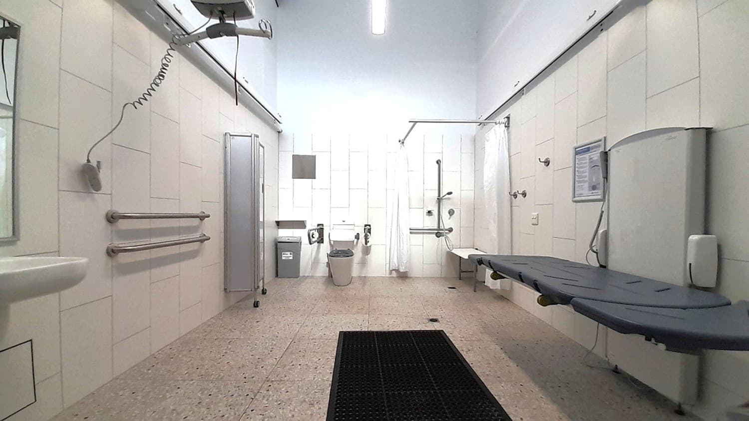 A spacious, accessible bathroom with a speckled floor and white walls. On the left, there are grab bars and a sink. In the centre, a toilet with support rails and a bin nearby. On the right, a shower area with a foldable seat and a long, adjustable bench. Overhead, a ceiling track hoist system is visible. The ceiling is plain with a fluorescent light fixture.