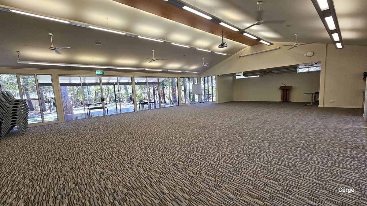 A spacious, empty hall with printed grey carpet. The pitched ceiling features rows of fluorescent lights and ceiling fans. A brown wooden table stands at the far end. Glass walls and glass doors cover the left side. Plastic chairs are stacked on the left.