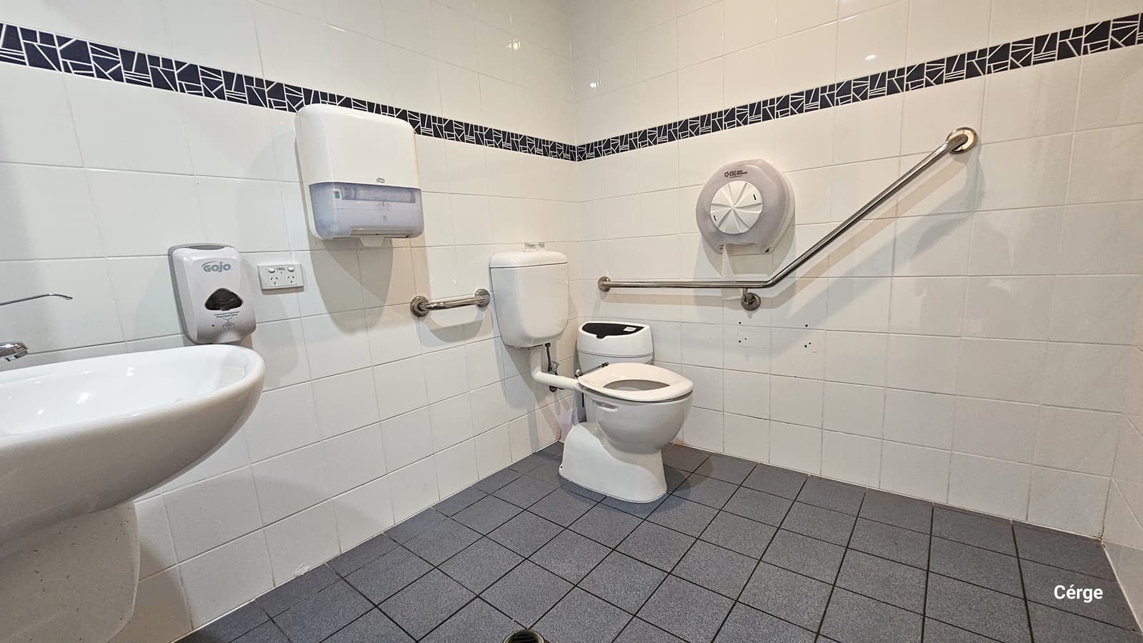 Inside the accessible toilet, a wash basin is mounted to the wall on the left, along with a soap and tissue dispenser. In the corner is a toilet bowl, a rubbish bin, and stainless hand grabs. The wall has white tiles with black printed accents, and the floor has grey tiles.