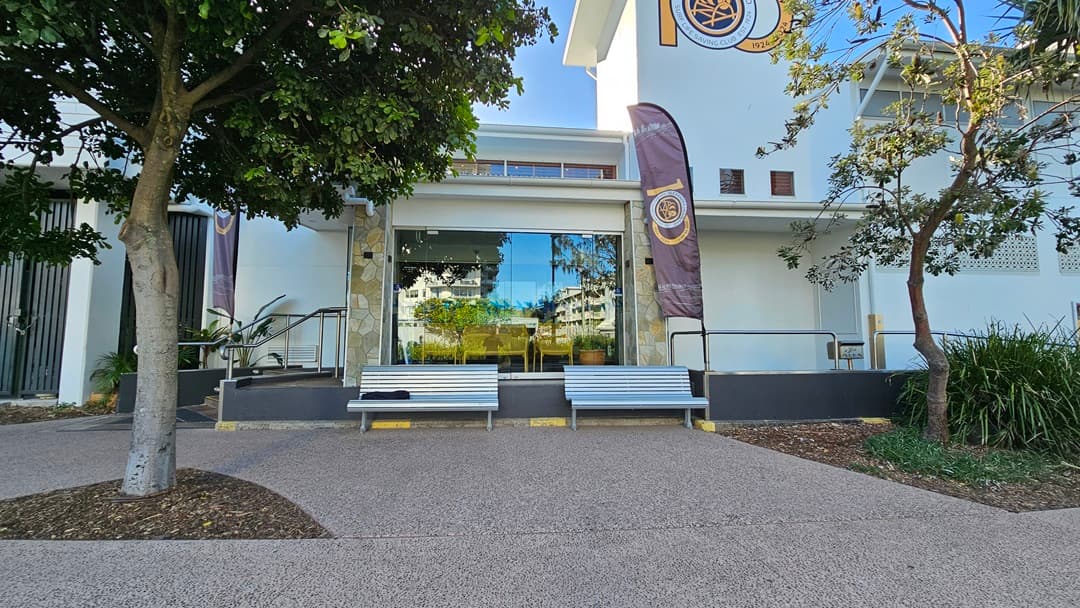 On the left, there is a tree with a thick trunk and green foliage. To the right, another tree with sparse leaves stands near a building. In the middle, a large glass window reflects the surrounding area, flanked by two grey benches with yellow accents. The background features a white building with a purple and gold flag. The floor is a textured, light brown pavement.