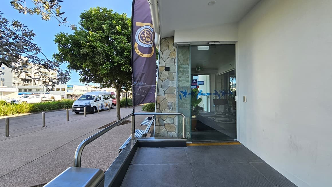 On the left, there is a tree with dense foliage next to a pathway where several vehicles are parked. In the middle, a vertical banner is attached to a pole near a stone wall. On the right, there is a glass door entrance with a metal handrail and a small bench beside it. The background includes more trees and buildings. The floor is a mix of concrete paving and a textured path, and the ceiling above the entrance is white with recessed lighting.