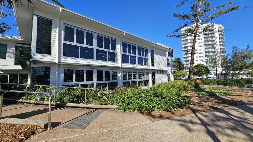 On the left, there is a white building with large windows and a metal handrail leading to a ramp. In the middle, there are bushes and plants in front of the building. On the right, a tall white apartment building is visible in the background, with trees and a pathway in the foreground. The floor is a mix of concrete paving and textured paths.