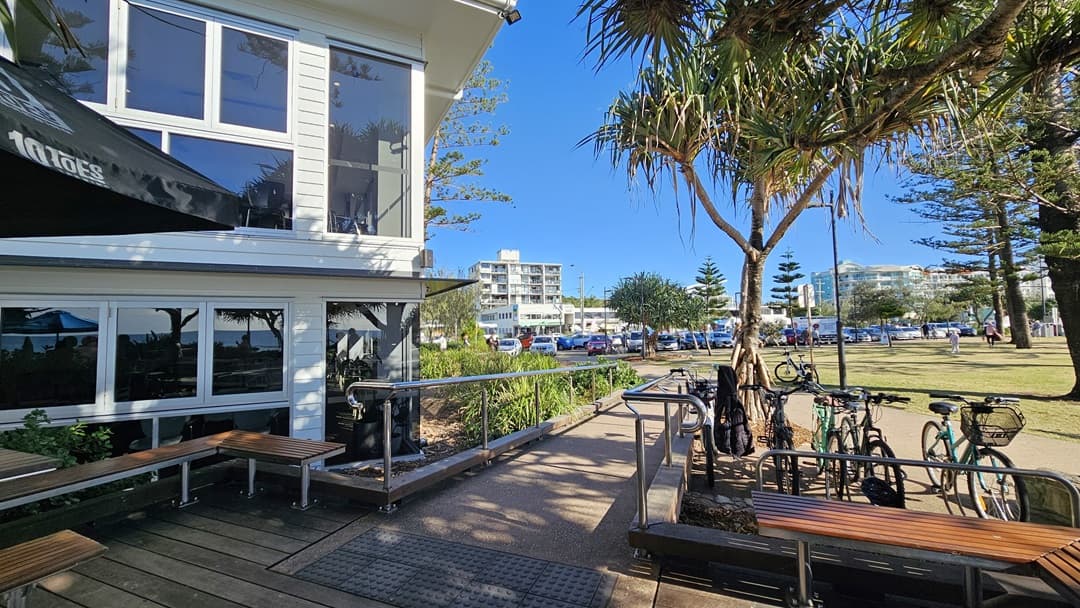 On the left, there is a white building with large windows and a black awning, next to a wooden bench and a metal handrail leading to a ramp. In the middle, there are several trees and a grassy area with parked bicycles. On the right, more trees and a pathway lead to a distant white apartment building. The foreground features a mix of wooden decking and concrete paving.