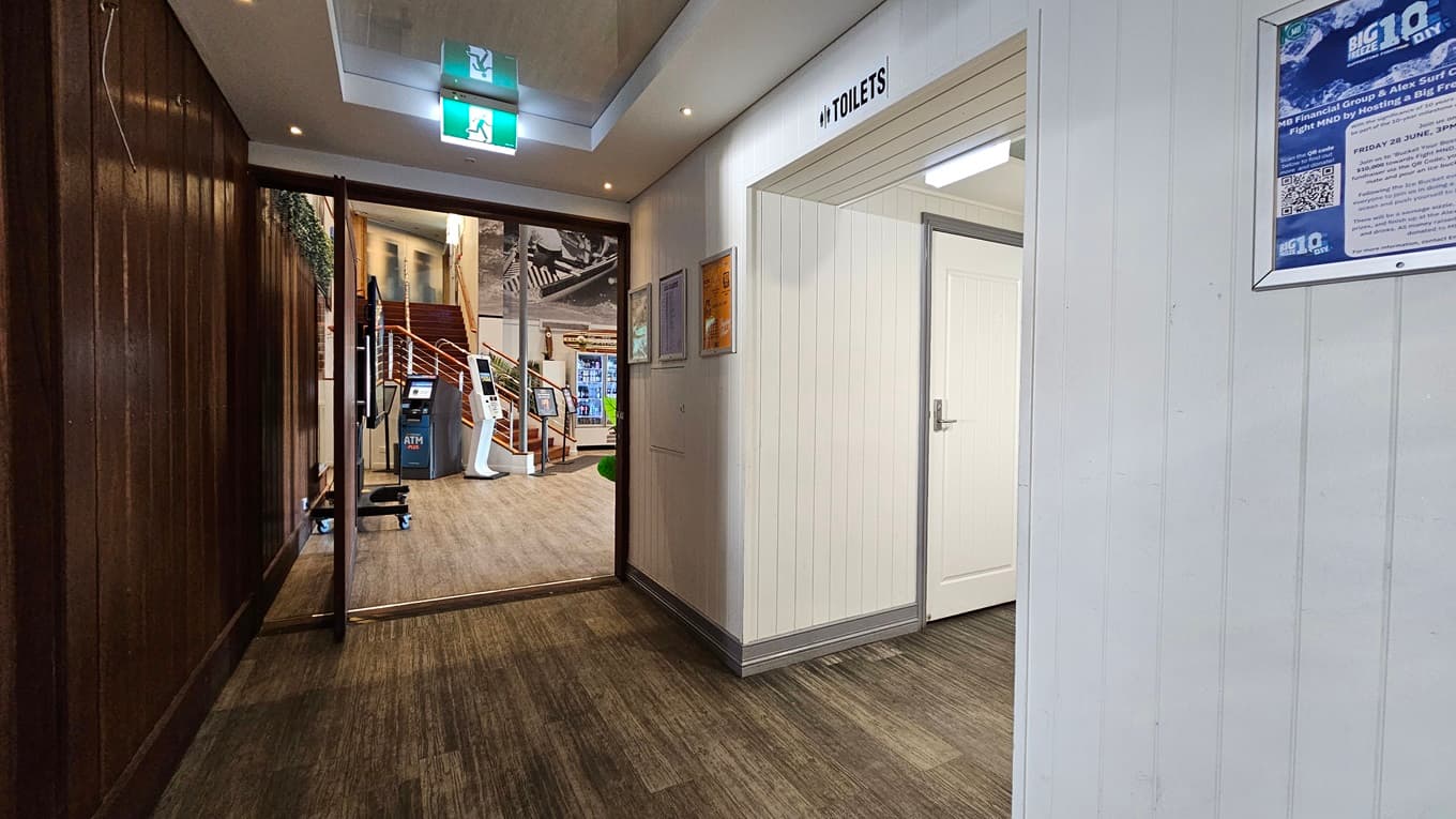 On the left, there is a wooden panelled wall leading to an open area with a staircase and a surfboard display. In the middle, there is a hallway with a wooden floor and a green exit sign on the ceiling. On the right, there is a white wall with a door labelled "Toilets" and a blue poster beside it. The floor is a mix of wooden and carpeted areas, and the ceiling has recessed lighting and an exit sign.
