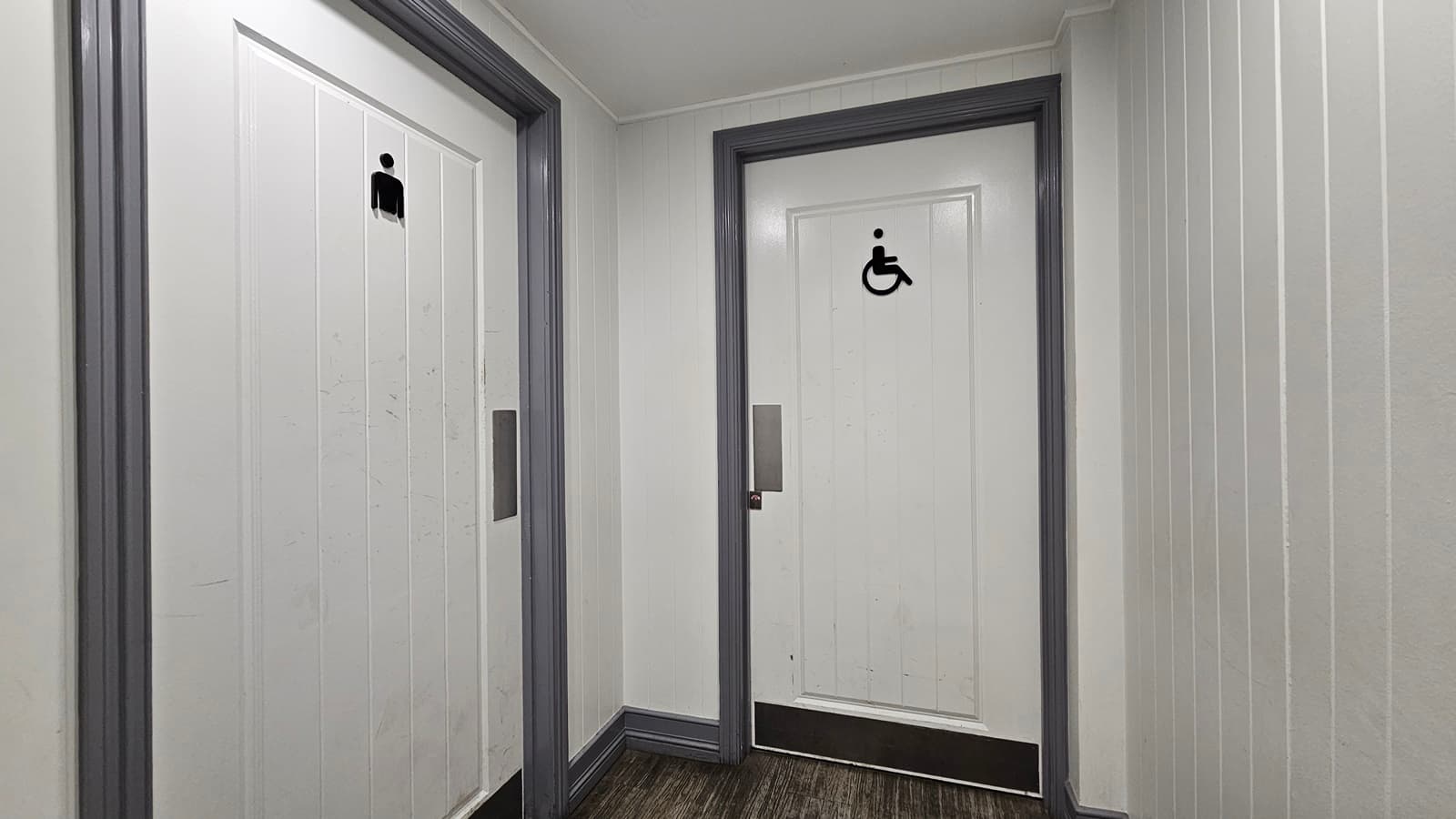 On the left, there is a white door with a male restroom symbol. In the middle, there is a white door with a wheelchair accessible symbol. Both doors have grey frames. On the right, there is a white panelled wall. The floor is dark wood, and the ceiling is white with recessed lighting.