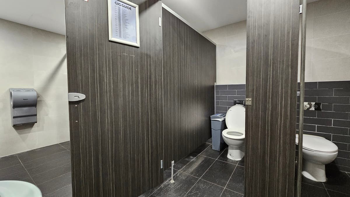 On the left, there is a grey paper towel dispenser mounted on a beige tiled wall. In the middle, there are two dark wood bathroom stalls, one with a closed door and the other open, revealing a white toilet and a blue bin. On the right, there is another white toilet with a grey tiled wall behind it. The floor is covered in dark grey tiles, and the ceiling is white with recessed lighting.