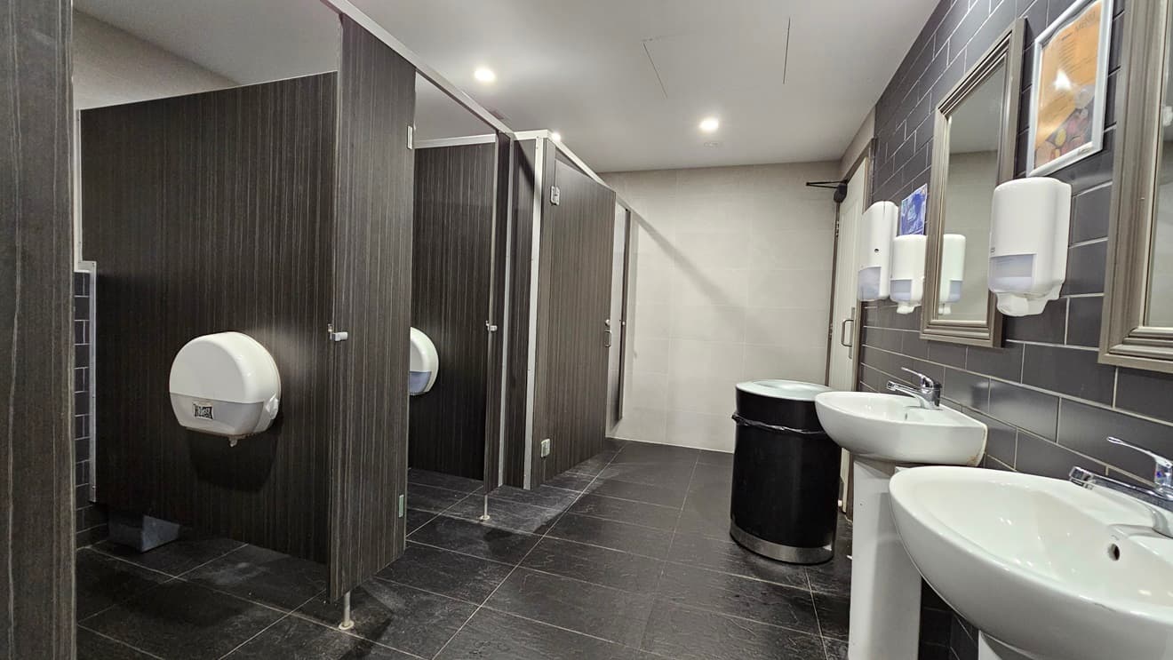 On the left, there are three dark wood bathroom stalls with closed doors, each with a white toilet paper dispenser. In the middle, there is a black bin against a beige tiled wall. On the right, there are two white sinks with mirrors above them, and three white soap dispensers mounted on a grey tiled wall. The floor is covered in dark grey tiles, and the ceiling is white with recessed lighting.