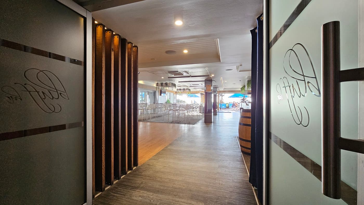 On the left, there is a frosted glass door with the word "Bluff" etched on it, partially open, revealing a wooden panelled wall. In the middle, there is a hallway with a wooden floor leading to a dining area with tables and chairs. In the background, there is a bar area with a wooden barrel and a blue umbrella visible through the windows. On the right, there is another frosted glass door with the word "Bluff" etched on it, also partially open. The ceiling is white with recessed lighting.