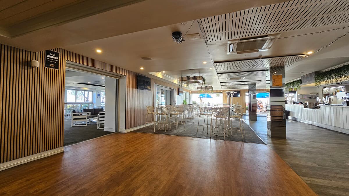 On the left, there is a wooden panelled wall with a "No Smoking" sign and an open doorway leading to a seating area with white chairs and tables. In the middle, there is a dining area with white tables and chairs on a dark carpet. In the background, there are large windows with views of the outside and a bar area with a blue umbrella. On the right, there is a long white bar counter with bottles and glasses. The floor transitions from wood to carpet to tile, and the ceiling has recessed lighting.