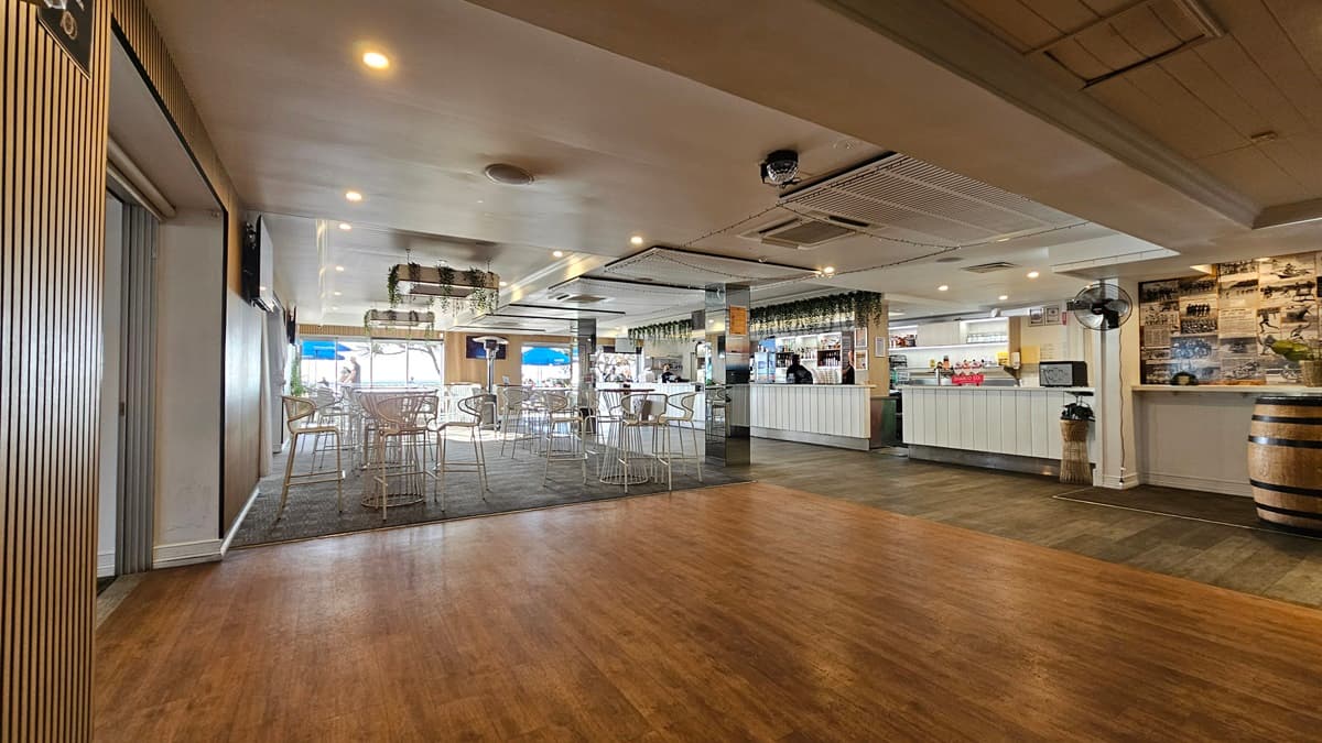 On the left, there is a wall with vertical blinds and a partially open sliding door. In the middle, there is a dining area with white metal chairs and tables on a dark carpet. In the background, there are large windows with views of the outside and blue umbrellas. On the right, there is a long white bar counter with bottles and glasses, a wooden barrel, and a wall with framed photos. The floor transitions from wood to carpet to tile, and the ceiling has recessed lighting.
