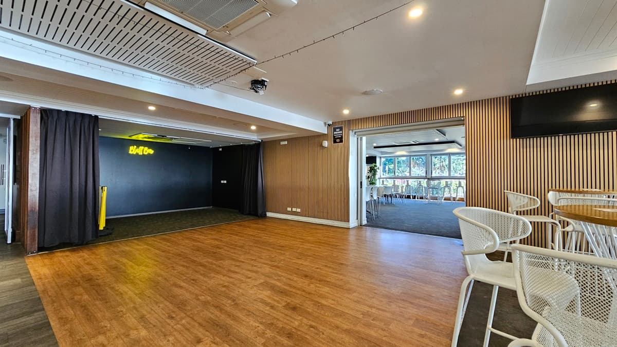 On the left, there is a black curtain partially covering a doorway with a neon "Bluff" sign above it. In the middle, there is an open space with a wooden floor leading to another room with large windows and white chairs. In the background, the room has a dark carpet and more white chairs. On the right, there is a wall-mounted TV above a wooden panelled wall and a seating area with white metal chairs and tables. The ceiling is white with recessed lighting and air conditioning units.