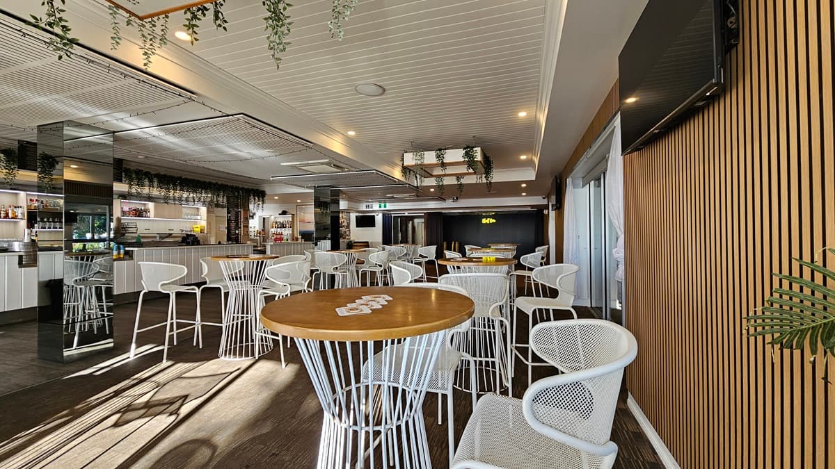 On the left, there are white cabinets and a bar area with shelves of bottles and glasses, decorated with hanging plants. In the middle, there are round wooden tables with white metal chairs on a dark carpet. In the background, there is a stage area with a black curtain and a neon "Bluff" sign. On the right, there is a wooden panelled wall with a wall-mounted TV and a sliding glass door. The floor is dark carpet, and the ceiling is white with recessed lighting and hanging plants.