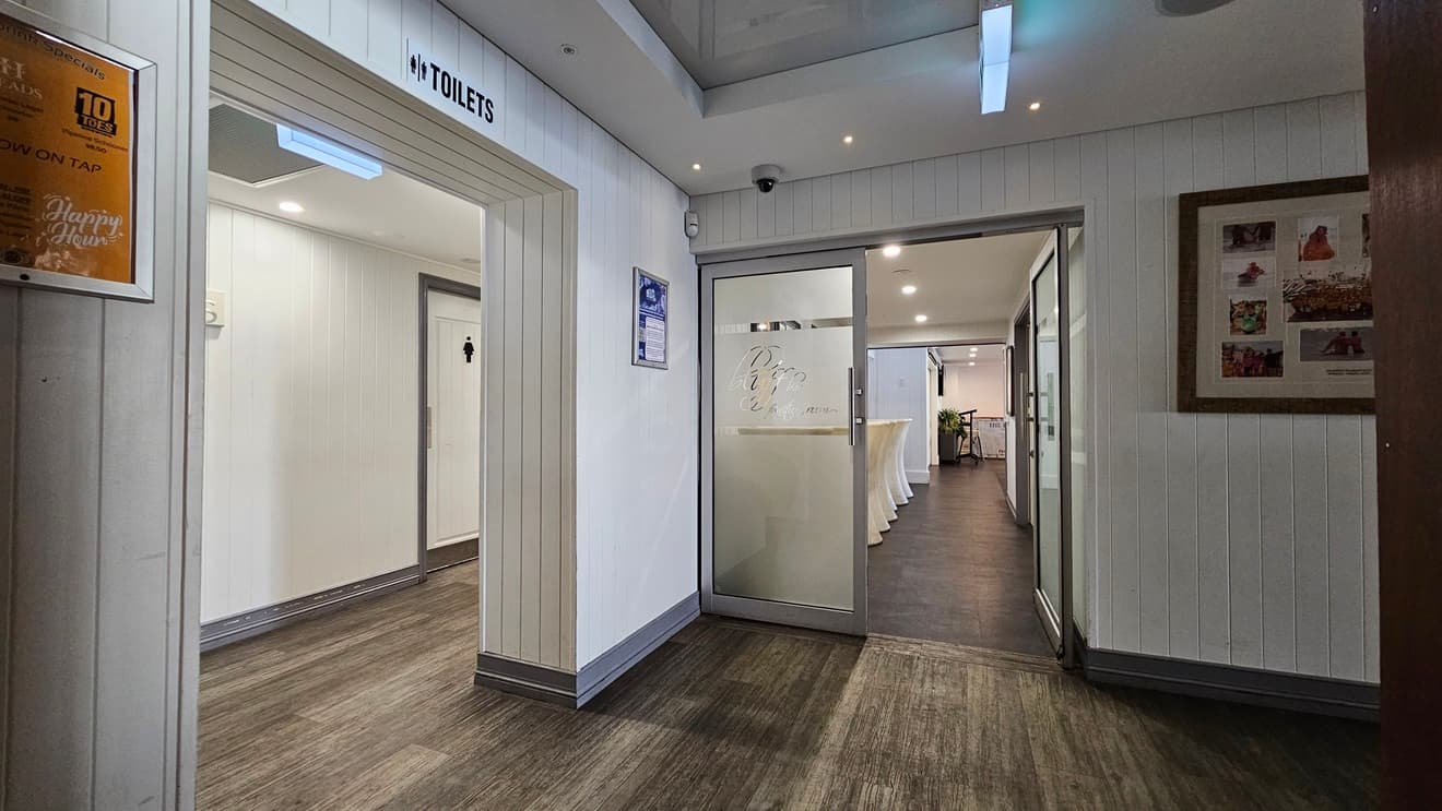 On the left, there is a doorway with a sign above it reading "Toilets," leading to a hallway with white doors. In the middle, there is a glass door with "Bluff" etched on it, opening into a room with white walls and a dark floor. In the background, there is a long table with a white tablecloth and a lit exit sign. On the right, there is a white panelled wall with a large framed collage of photos. The floor is dark wood, and the ceiling is white with recessed lighting.