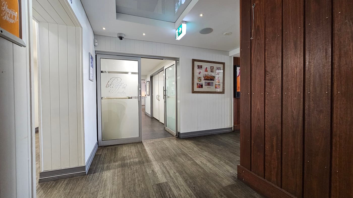 On the left, there is a white panelled wall with a small framed picture and a doorway leading to a hallway. In the middle, there is a glass door with the word "Bluff" etched on it, opening into another room with white walls and dark flooring. In the background, there are more glass doors and a lit exit sign. On the right, there is a wooden panelled wall with a large framed collage of photos. The floor is a mix of dark and light wood, and the ceiling is white with recessed lighting.