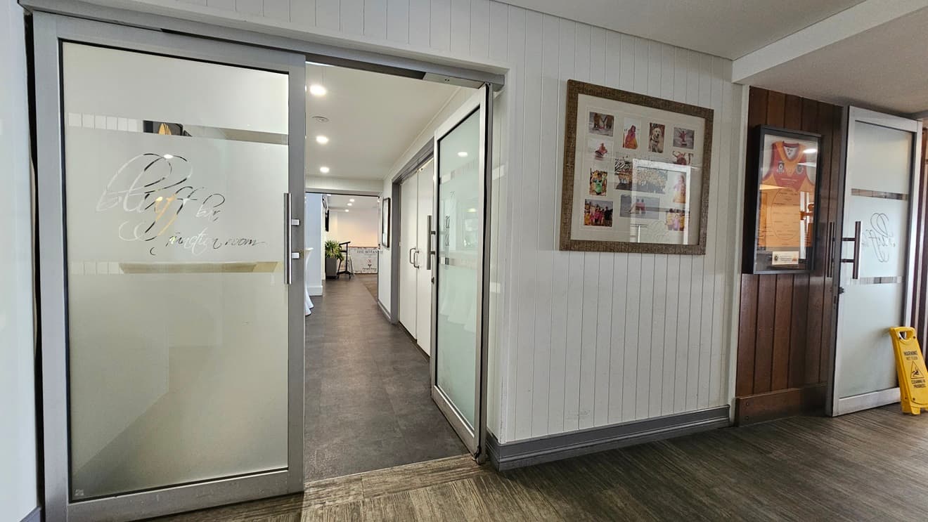 On the left, there is a glass door with "Bluff" etched on it, leading to a hallway with white walls and dark flooring. In the middle, there is another glass door with the same etching, also leading to the hallway.The hallway extends to a well-lit area with white cabinets. On the right, there is a white panelled wall with a large framed collage of photos and a wooden panelled section with a bulletin board. The floor has timber and the ceiling is white with recessed lighting.